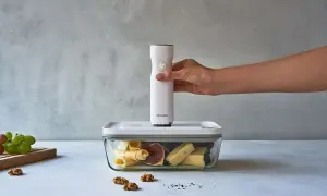 Ręka trzyma białą pompkę do soku nad szklanym pojemnikiem z serem, figami i winogronami.
