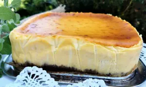 Pyszny sernik z mleka w proszku, z lekko przypieczoną skórką, na metalowej tacy, otoczony zielenią i białą koronką.
