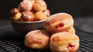 Puszyste ciasto na pączki, posypane cukrem, z widocznym nadzieniem. W tle miska z mniejszymi pączkami w polewach.