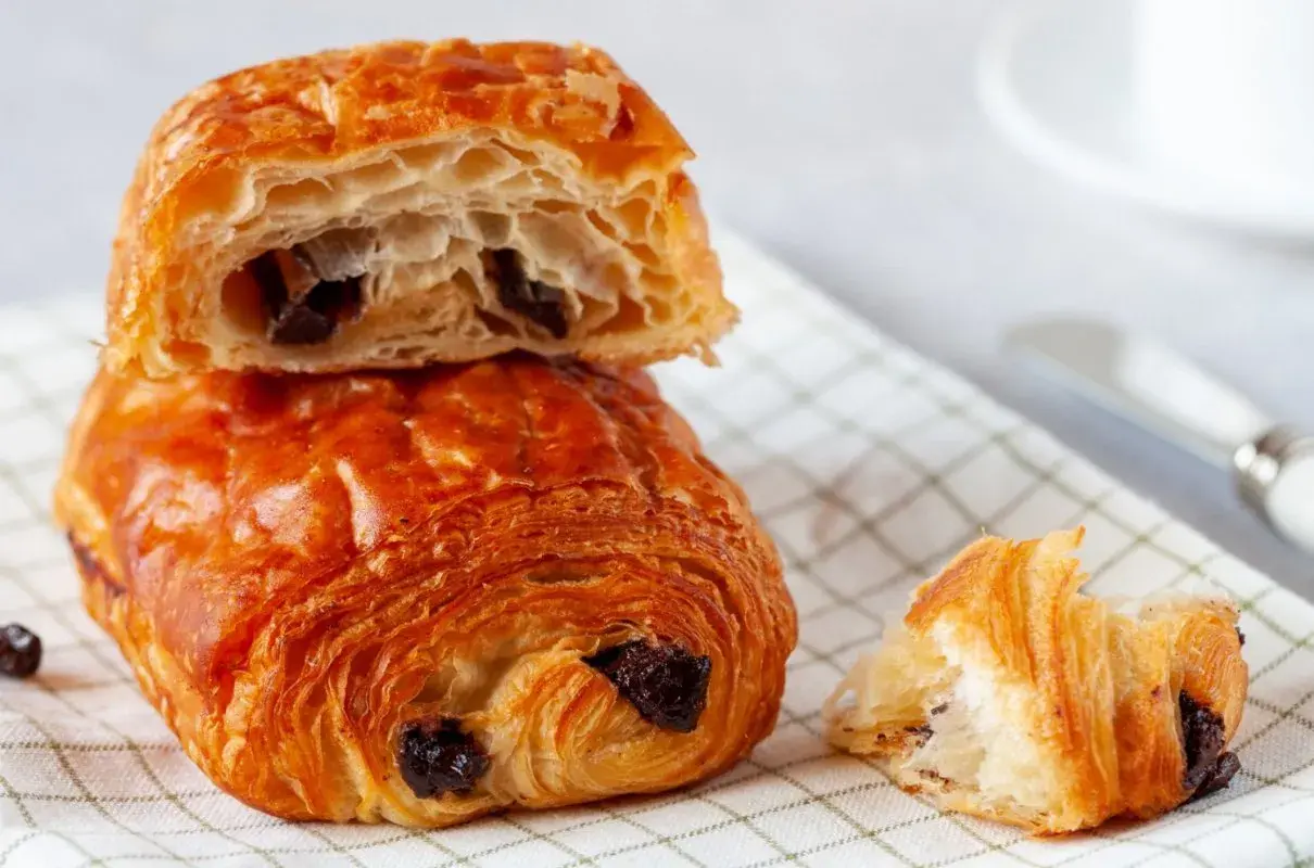 Chrupiące, złociste pain au chocolat z kawałkami czekolady, jeden przekrojony, ukazujący puszyste wnętrze.