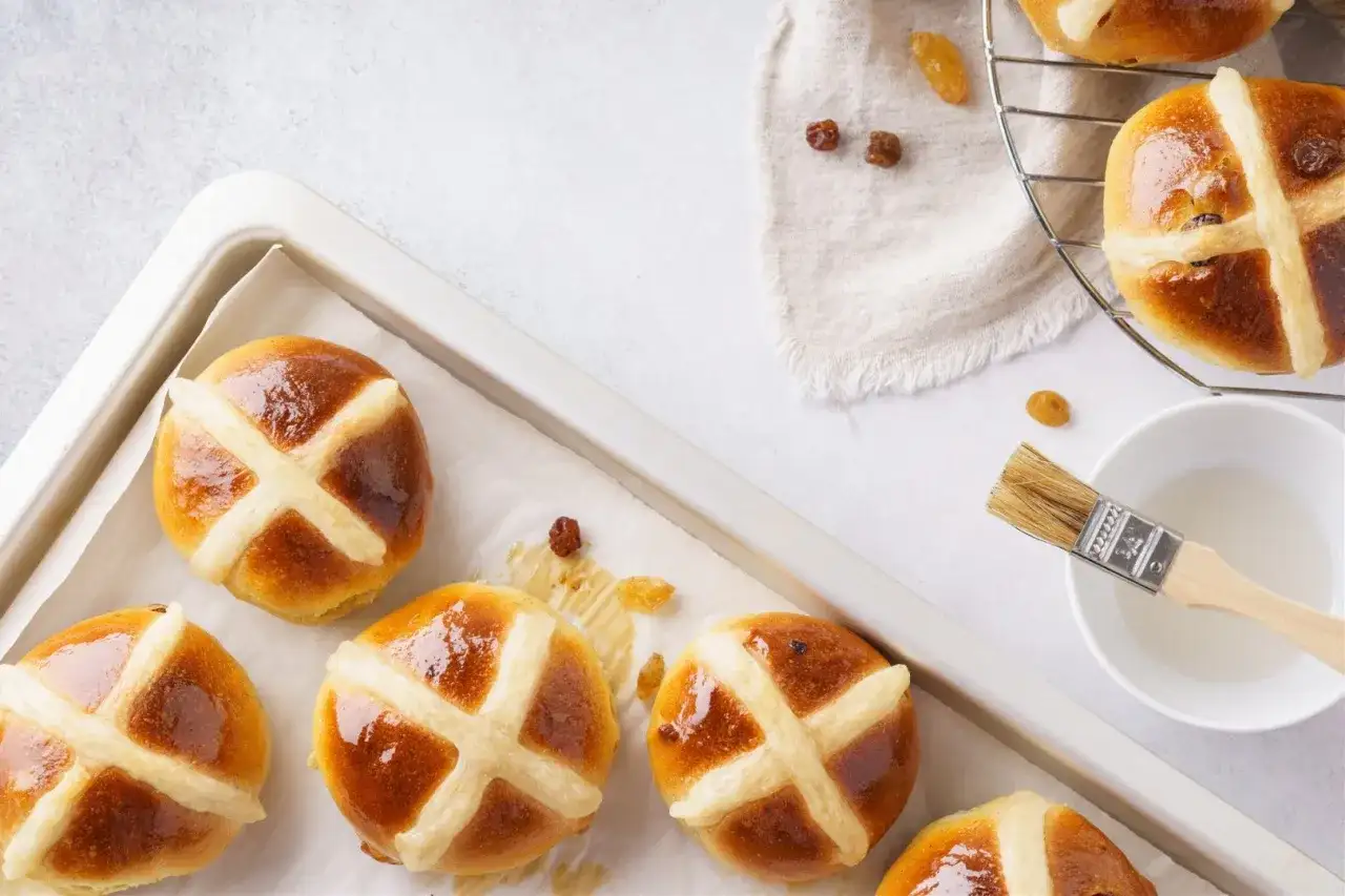 Aksamitne, błyszczące hot cross buns z rodzynkami, gotowe do pieczenia. Pędzelek i lukier czekają.