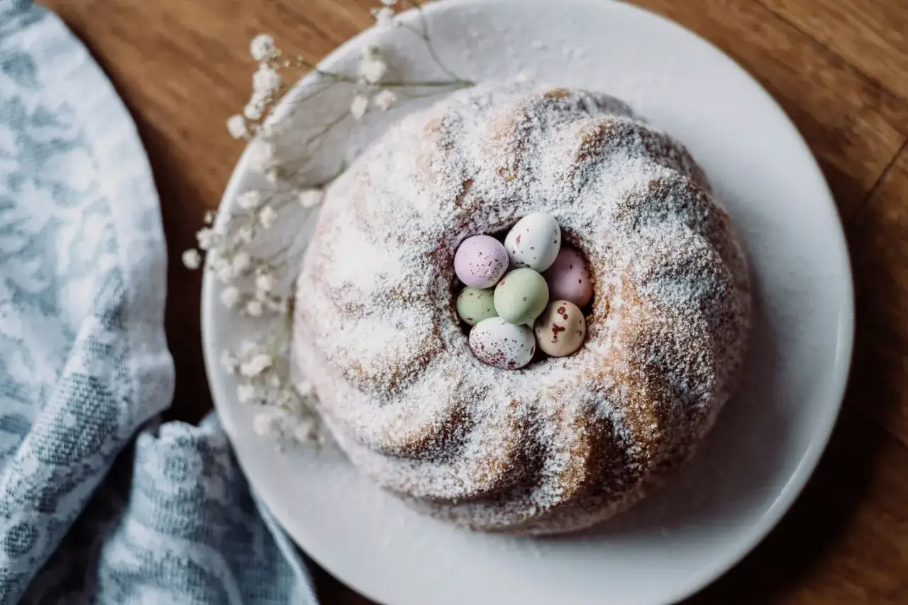 Babka wielkanocna przepis: puszysta babka z cukrem pudrem i kolorowymi jajeczkami w środku, ozdobiona drobnymi białymi kwiatuszkami.