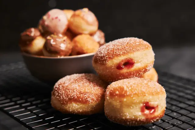 Puszyste ciasto na pączki, posypane cukrem, z widocznym nadzieniem. W tle miska z mniejszymi pączkami w polewach.